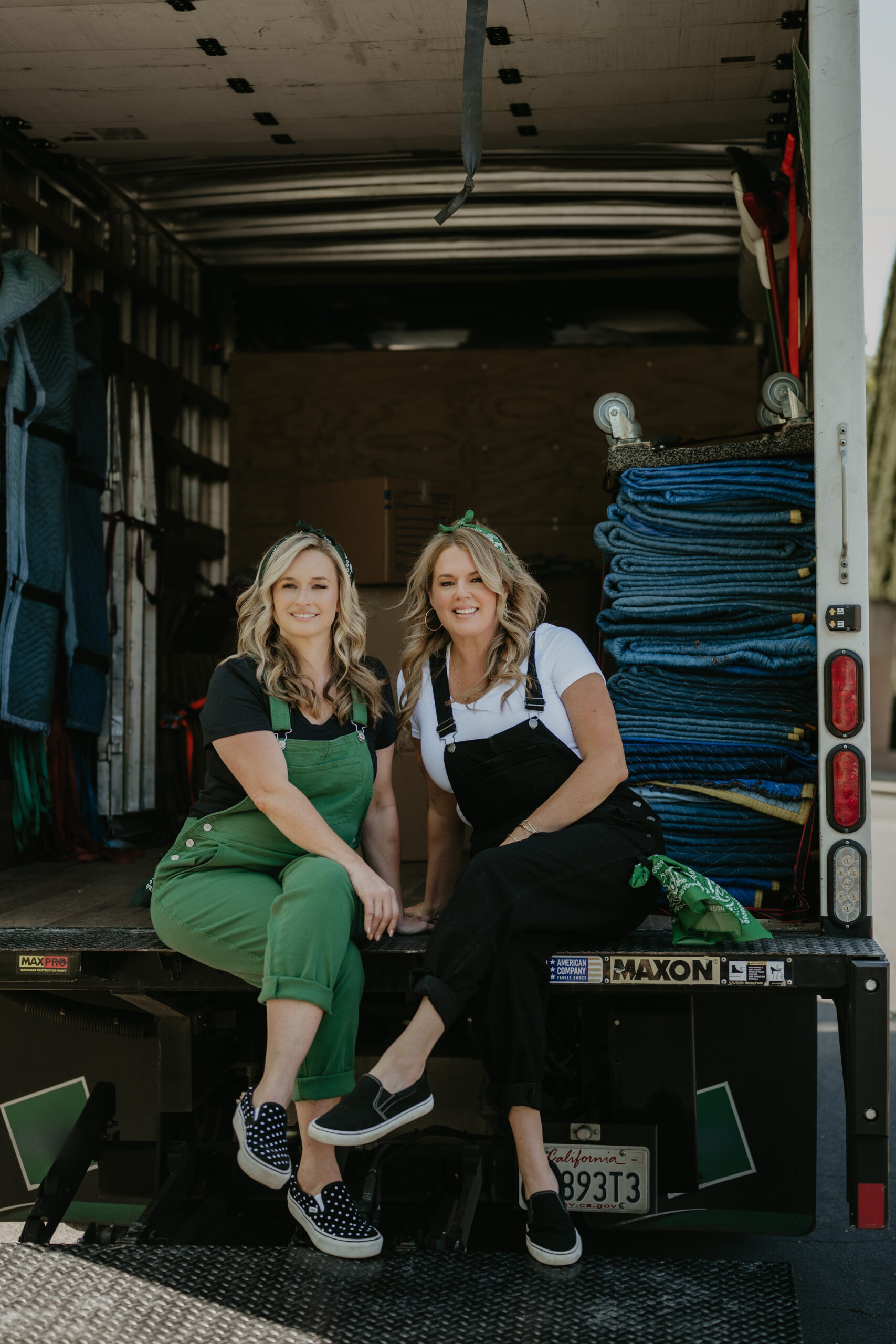 4829cd18-0ac0-4e2d-be1e-ed01731d7384 2 blonde women in overalls sitting in bvack of moving truck