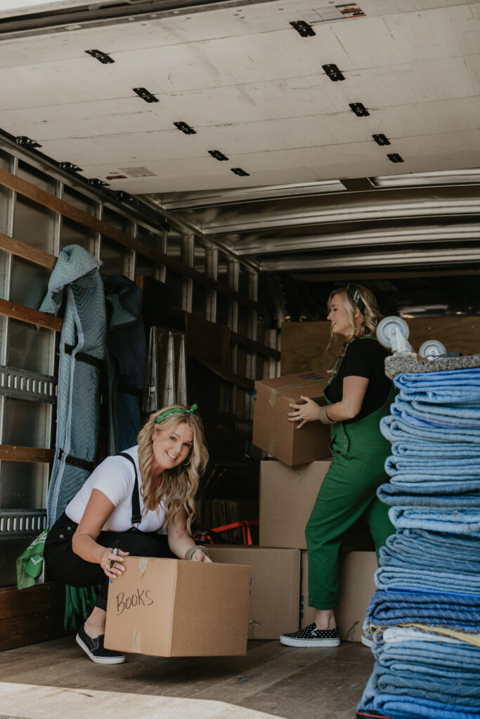 Two women from Betsy’s Moving loading boxes into a moving truck, surrounded by moving pads and labeled boxes.